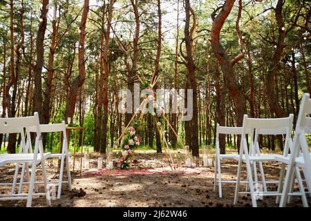 Wunderschöne böhmische Tipi-Bogendekoration auf der Hochzeitsfeier im Freien im Pinienwald mit Kegeln. Stühle, floristische Blumenkompositionen aus Rosen Stockfoto