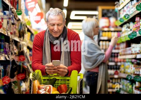 Älterer Mann, der die Einkaufsliste auf dem Smartphone überprüft und mit seiner Frau kauft Stockfoto