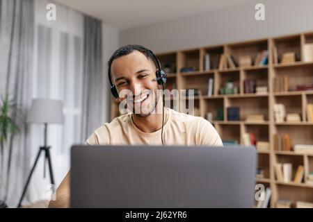 Fröhlicher Mann aus dem Nahen Osten, der über einen Laptop mit kabellosen Kopfhörern Video-Chat führt, auf den Bildschirm blickt und lächelt Stockfoto