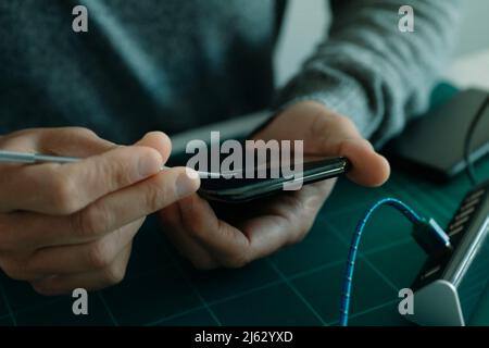 Nahaufnahme eines jungen kaukasischen Mannes, der den zerbrochenen Bildschirmschutz eines Smartphones entfernt und an einem Tisch sitzt Stockfoto