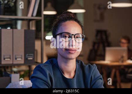 Porträt einer lächelnden attraktiven jungen Geschäftsfrau aus dem biracial Bereich in einer Brille, die im Büro an den Regalen sitzt Stockfoto