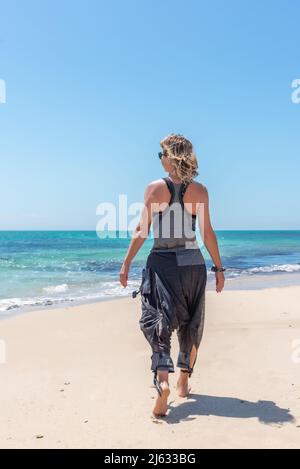 Kaukasische blonde Frau, die an einem sonnigen Tag in lässiger Kleidung an einem goldenen Sandstrand entlang läuft Stockfoto