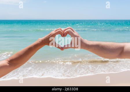 Hände bilden ein Herz vor einem Strand aus goldenem Sand und türkisfarbenem Wasser. Liebe im Paradies Stockfoto