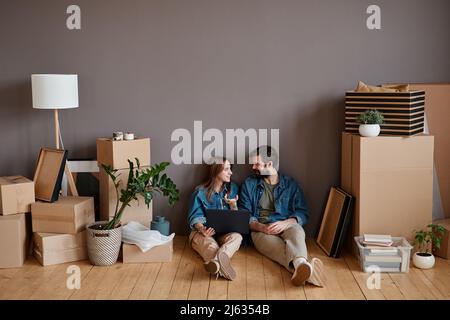 Junges Paar, das in einer neuen Wohnung sitzt und auf dem Laptop Internet surft, um nach Innenarchitektur-Bildern zu suchen und eines zu wählen Stockfoto