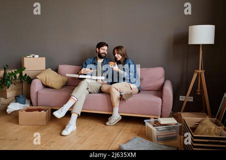 Junge kaukasische Mann und Frau entspannen zusammen auf dem Sofa essen köstliche Pizza im Loft Wohnzimmer nach dem Umzug in eine neue Wohnung Stockfoto