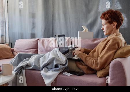 Horizontale Aufnahme einer erwachsenen kaukasischen Frau mit Erkältung, die zu Hause eine SMS an einen Laptop geschickt hat Stockfoto