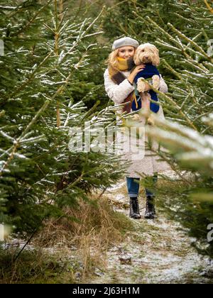 Junge blonde Frau mit einem Hund, der zwischen schneebedeckten Weihnachtsbäumen steht. Stockfoto