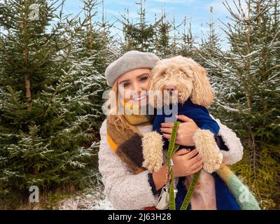 Junge kaukasische Frau, die in einem warmen Hut und Mantel gekleidet ist und einen Hund hält, der vor schneebedeckten Weihnachtsbäumen posiert. Stockfoto