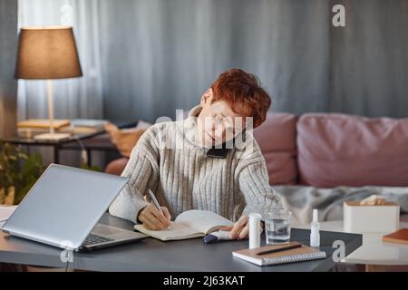 Reife Frau mit Grippe sitzt im Wohnzimmer zu Hause am Schreibtisch und telefoniert mit Kollegen und macht sich Notizen Stockfoto