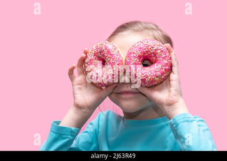 Ein niedliches lustiges Mädchen in einem blauen T-Shirt hält leuchtend rosa Donuts wie eine Brille in der Nähe ihrer Augen und sieht dich auf einem rosa Hintergrund an. Fröhliche Kindheit Konz Stockfoto