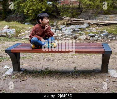 Einsamer nachdenklicher Latino kleiner Junge, der auf einer Eisen- und Holzbank neben einem Bach in städtischen Kleidern sitzt und die Landschaft beobachtet. Stockfoto