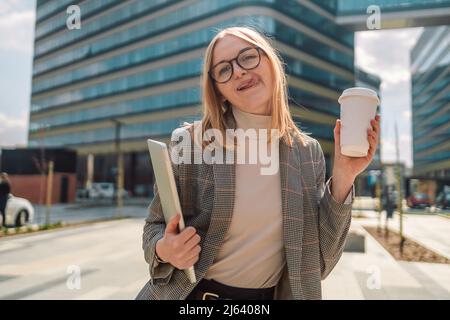 Europäische blonde attraktive Geschäftsfrau in trendigen Kleidern mit einem Laptop und Take Away Bio-Papier-Kaffeetasse, zeigt Zunge, posiert draußen. Amerikanisch Stockfoto