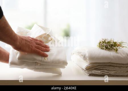 Mann, der frisch gewaschene und gebügelte saubere Hemden auf einem weißen Tisch und Fensterhintergrund in einem Zimmer zurückließ. Vorderansicht. Horizontale Zusammensetzung. Stockfoto