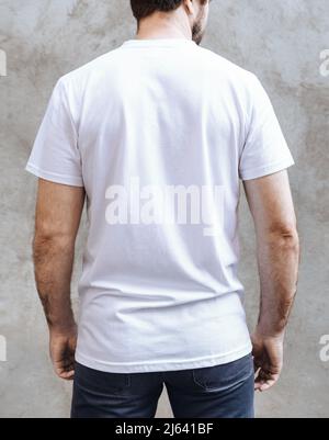 Junger Mann in einem leeren weißen legeren T-Shirt. Rückansicht auf einem Hintergrund hellgrauer Betonwände. Design und Layout des Herren-T-Shirts zum Bedrucken. Stockfoto