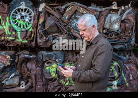 Ein Mann, der sein Mobiltelefon überprüft, während er vor einer Wand aus zerquetschten Autos steht. Ironischerweise haben die Fahrer dieser Autos vielleicht das gleiche getan. Stockfoto