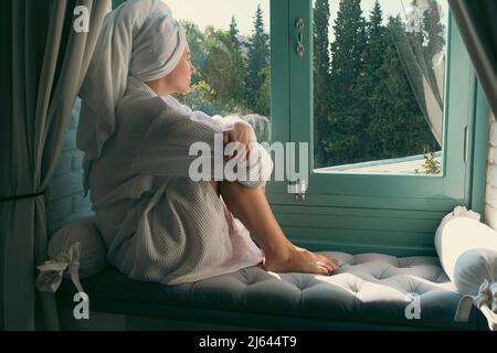 Frau, die nach dem Bad im Bademantel auf der Fensterbank sitzt. Relax und Spa-Konzept. Gesundheit der Frauen Stockfoto