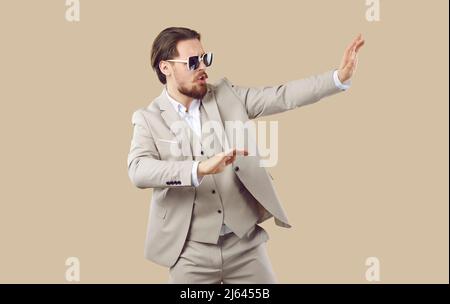 Lustiger junger Mann im klassischen Anzug und coole Sonnenbrillen tanzen auf beigem Studiohintergrund Stockfoto