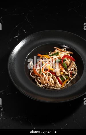 Japanische udon-Nudeln in einem Wok gebraten. Udon-Nudeln mit Gemüse auf dunklem Hintergrund. Asiatische Küche, Wok-Menü. Stockfoto