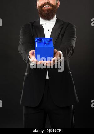 Fröhlicher Mann in Smoking Fliege auf schwarzem Hintergrund präsentiert Geschäft Belohnung Box. Anlass Gruß Stockfoto