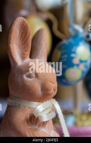 Nahaufnahme einer kleinen Terrakotta-Osterhasen-Figur mit hellgelbem Band vor dekorierten Ostereiern-Dekorationen. Stockfoto