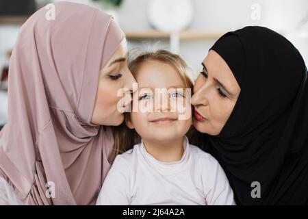 Muslimische Tochter und glückliche ältere Großmutter küssen Kind Enkelin auf den Wangen gratuliert kleinen Mädchen mit Geburtstag Ausdruck von Sorgfalt und Liebe, drei Generationen von Frauen Konzept Stockfoto