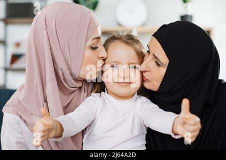 Muslimische Tochter und glückliche ältere Großmutter küssen Kind Enkelin auf den Wangen gratuliert kleinen Mädchen mit Geburtstag Ausdruck von Sorgfalt und Liebe, drei Generationen von Frauen Konzept Stockfoto