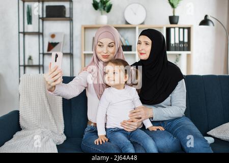 Portrait of Overjoyed drei Generationen muslimischer Frauen haben Spaß zu Hause Machen Sie gemeinsam Selfie, lächelndes kleines Mädchen mit junger arabischer Mutter und älterer Großmutter Machen Sie ein Selbstporträt Stockfoto