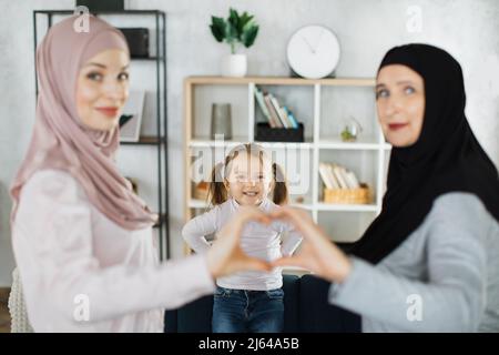 Portrait of happy drei Generationen von islamischen Frauen posieren zusammen zeigen Liebe Herz Hand Geste. Lächelndes, kleines muslimisches Mädchen, Kind mit junger Mutter und älterer Großmutter, fühlt sich vereint. Stockfoto