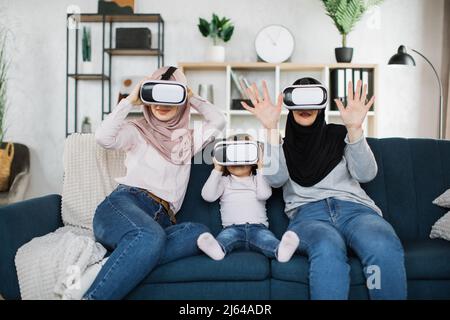 Glückliche drei Generationen muslimischer Frauen, die in modernen Augmented-VR-Headset-Reality-Brillen im Wohnzimmer zu Hause zusehen. Virtual Reality und futuristische Technologie, Ruhestand und Freizeit Konzept Stockfoto