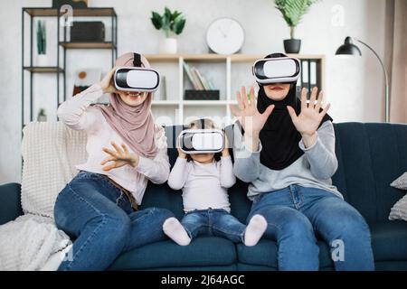 Glückliche drei Generationen muslimischer Frauen, die in modernen Augmented-VR-Headset-Reality-Brillen im Wohnzimmer zu Hause zusehen. Virtual Reality und futuristische Technologie, Ruhestand und Freizeit Konzept Stockfoto
