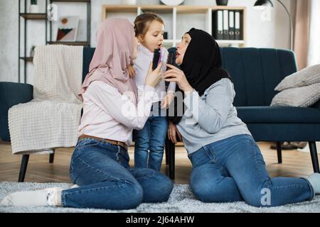 Drei Generationen muslimischer Frauen sitzen auf den Knien auf dem Boden in der Nähe des Sofas im Wohnzimmer zu Hause und singen ihre Lieblingslieder mit einem Kamm anstelle eines Mikrofons. Stockfoto