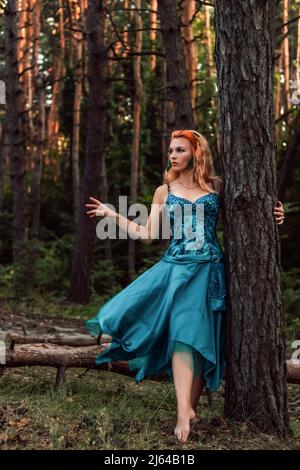 Ein schönes Mädchen mit roten Haaren steht in der Nähe eines Baumes im Wald und schaut weg Stockfoto