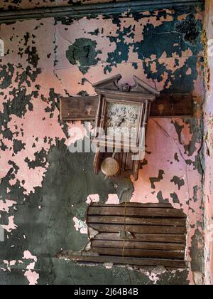 Eine krumme Uhr und eine alte Gipswand, die die Farbschichten zeigt, sind die Armaturen in diesem verlassenen Haus. Stockfoto