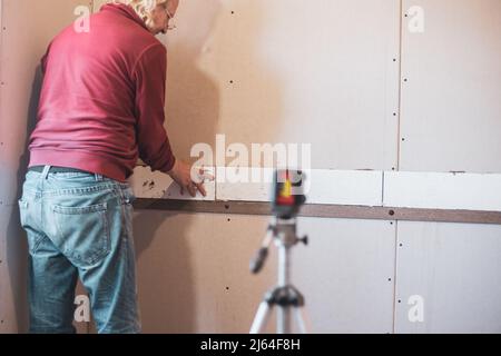 Ein Mann legt keramische Fliesen auf Fliesenkleber an einer Wand mit einem Laser-Level. Hausrenovierung, Flieseninstallation in der Küche. Stockfoto