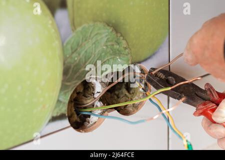 Ein Mann verbindet elektrische Leitungen, um eine Steckdose in einer gefliesten Wand zu installieren. Renovierung und Verbesserung des Hauses. Stockfoto