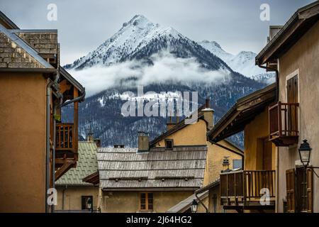 Briancon, Frankreich - 15 Mar 2022: Schneebedeckter Serre Chevalier-Gipfel, eingerahmt von den Häusern des Skigebiets; schönste Straßentapete Stockfoto