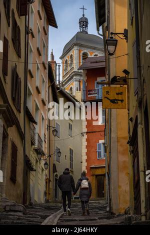 Briancon, Frankreich - 15 Mar 2022: Junges Paar, das in einer farbenfrohen Straße von Briancon die Treppe hochgeht und sich die Hände hält Stockfoto