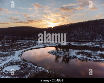 Winteransicht auf Flusskurve mit szenischer Reflexion. Region Smijewski auf dem Fluss Siwerskyi Donez in der Ukraine. Sonnenuntergangssonne in Wolken über verschneiten Wald Stockfoto