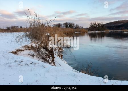 Winterblick auf den Fluss mit malerischen lila Wolken mit Spiegelung im spiegelnden Wasser und trockenem Schilfgras am Ufer. Wilde Landschaft mit Schnee bedeckt Stockfoto