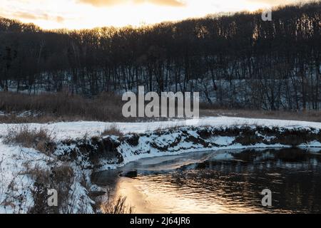 Blick auf den Winter am gefrorenen Flussufer. Der Fluss Siwerskyi Donez ist in der Ukraine schneebedeckt. Sonnenuntergangssonne in Wolken über einem bewaldeten Hügel Stockfoto