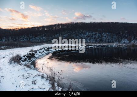 Winteransicht auf gefrorener Flusskurve mit szenischer Spiegelung der Wolken. Der Fluss Siwerskyi Donez ist in der Ukraine schneebedeckt. Sonnenuntergangswolken über einem bewaldeten Hügel Stockfoto