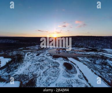 Winterüberblick auf verschneiten Fluss. Region Smijewski auf dem Fluss Siwerskyi Donez in der Ukraine. Die Sonnenuntergangssonne scheint über einem bewaldeten Hügel Stockfoto