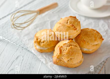 Selbstgemachte Profiterolen oder Eklare auf einem Metallgitter. Daneben befindet sich ein Schneebesen, dahinter steht eine Tasse auf einer Untertasse. Traditionelles französisches Dessert. Weiß Stockfoto