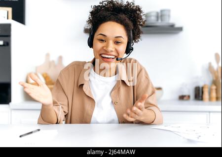 Hübsches afroamerikanisches stilvolles Mädchen mit Headset, Mentor oder Student, führt ein Online-Meeting mit Kollegen oder Studenten durch, hält eine Rede über Unternehmensideen und -Pläne, lächelt. Online-Unterrichtskonzept Stockfoto