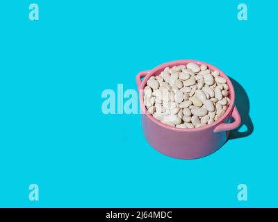 Getrocknete weiße Bohnen in einer rosa Schale isoliert auf einem blauen Hintergrund mit einem tiefen Schatten. Nahaufnahme, Kopierbereich. Stockfoto