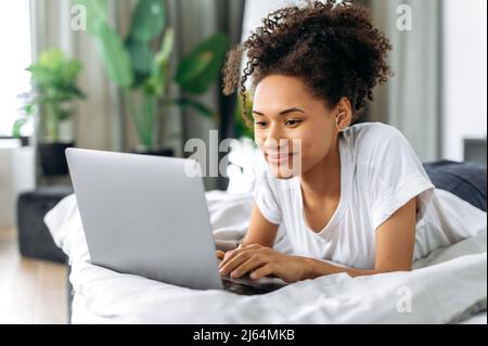 Ein nettes, positives afroamerikanisches Mädchen liegt zu Hause auf einem Bett auf dem Bauch, benutzt einen Laptop, durchsucht das Internet, chattet in sozialen Netzwerken, schaut sich ein Online-Webinar an, schreibt E-Mails und lächelt Stockfoto