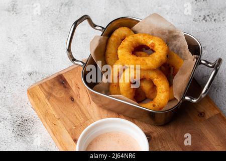 Deep - frittierte Zwiebelringe in einem Metallkorb mit weißer Sauce auf einem Holzbrett auf Marmorboden Stockfoto