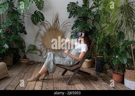 Aufgeregte junge Frau, die sich auf einen Stuhl lehnt und ein vr-Headset trägt, spielt in virtuellen Spielen im Heimgarten Stockfoto