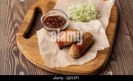 Verzehrfertige Schweine Würste in Speck auf Holzbrett gewickelt. Gebratene, herzhafte Würste in Speck verpackt, serviert mit Zwiebel und Sauce auf Holzboden Stockfoto