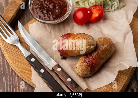 Verzehrfertige Schweine Würste in Speck auf Holzbrett gewickelt. Gebratene, herzhafte Würste in Speck verpackt, serviert mit Zwiebel und Sauce auf Holzboden Stockfoto